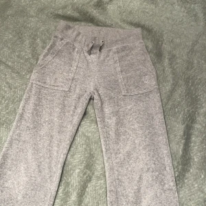 Grå mjukisbyxor från Juicy Couture - Säljer ett par grå mjukisbyxor från Juicy Couture i storlek XXS. Byxorna har en bekväm passform med resår och snörning i midjan. De har framfickor och bakfickor med knappar samt en broderad logga på ena benet. Byxorna är uppsydda och är därför kortare i benen än vad de var när de köptes. De är nu 75 cm vid inre benet.