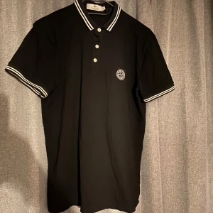 Svart pikétröja från Stone Island - Snygg svart pikétröja från Stone Island med storleken M men skulle säga att den sitter lika bra för S och broderad logga på bröstet. Tröjan har vita detaljer på krage och ärmslut samt knappar framtill. Perfekt för en stilren look.