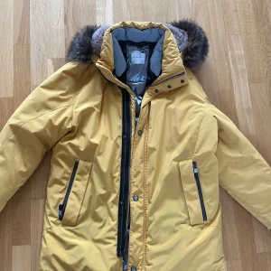 Gul vinterjacka/Parka - Snygg gul vinterjacka från Bugatti med avtagbar pälskant på huvan. Jackan har dragkedja och knappar framtill samt praktiska fickor med dragkedja. Storlek 50, helt ny och oanvänd!