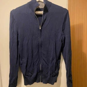 Mörkblå tröja med dragkedja - Säljer en stilren mörkblå tröja från Pierone med dragkedja och ribbad krage. Perfekt för en avslappnad look. Tröjan har långa ärmar och ribbade muddar vid ärmslut och nederkant. Tröjan ser lite trasslig ut, men strycks den är den så gott som ny 