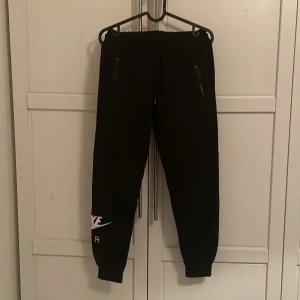 Svarta mjukisbyxor från Nike - Säljer ett par svarta mjukisbyxor från Nike med elastisk midja och snörning. Byxorna har dragkedjefickor och en vit Nike-logga på benet. Perfekta för en avslappnad stil.