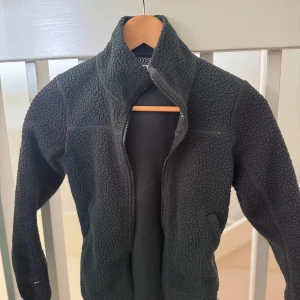 Grön fleecejacka/tröja - Mysig grön fleecejacka med dragkedja framtill. Perfekt för kyliga dagar med sin varma och mjuka känsla. Jackan har en hög krage och långa ärmar, vilket ger extra skydd mot kylan. En stilren och praktisk jacka för vardagsbruk.