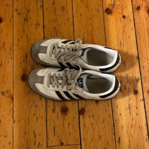 Adidas sneakers med svarta ränder - Klassiska Adidas sneakers i vitt med svarta ränder och grå detaljer. Skorna har snörning och en retro design som passar perfekt för en avslappnad stil.