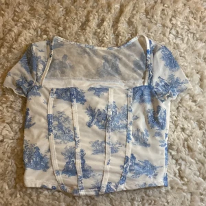 Vit och blå korsettopp liknande. Toppen passar lika bra för XS också. Eftersom att den kommer från shein så lägger jag priset på 30 kr. Har använts knappt.  - Säljer en söt vit och blå korsettopp med ett vackert mönster. Toppen har korta ärmar och en elegant design som passar perfekt för sommarens alla tillfällen.