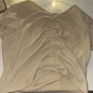 Beige ribbad T-shirt  från Lindex - Säljer en beige ribbad T-shirt från Lindex med korta ärmar och snörning framtill. Perfekt för en stilren look. Passar bra till både jeans och kjol.