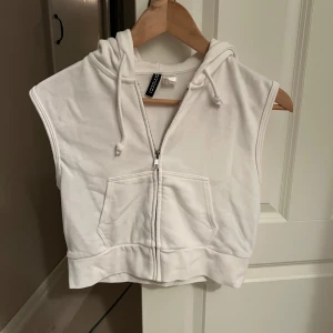 Vit croppad väst med dragkedja - Snygg vit croppad väst från HM med dragkedja och huva. Perfekt för en avslappnad stil. Västen har en praktisk ficka framtill och är ärmlös.