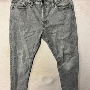 Grå jeans från Jack & Jones - Snygga grå jeans från Jack & Jones med en tapered passform. Stl. 28/30. Perfekta för en stilren look. De har en klassisk design med fem fickor och knappgylf.