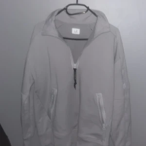 Grå tröja från C.P. Company - Snygg grå tröja från C.P. Company med dragkedja och hög krage. Tröjan har långa ärmar och praktiska fickor med dragkedja. Perfekt för en stilren och avslappnad look.