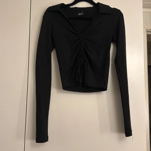 Svart ribbad topp från Gina Tricot - Snygg svart ribbad topp från Gina Tricot med långa ärmar och en stilren krage. Toppen har en knytning framtill som ger en fin detalj och en croppad passform!💓☺️