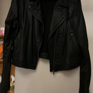 Svart bomulls jacka med nitar - Snygg svart bomull med coola nitar och asymmetrisk dragkedja. Perfekt för en edgy look. Jackan har långa ärmar och en klassisk bikerstil.