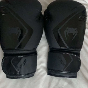 12oz boxningshandskar från Venum - Oanvända Snygga svarta boxningshandskar från Venum med en stilren design och logotyp på ovansidan. Handskarna har breda kardborreband för säker passform och extra vaddering för komfort och skydd under träning.