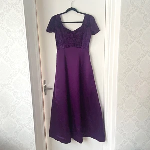 Lila klänning med mönstrad överdel - Elegant lila klänning med kortärmad design och v-ringad hals. Överdelen har ett vackert mönster som ger en lyxig känsla. Perfekt för speciella tillfällen.