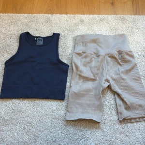 Mörkblå ribbad linnetopp och beiga ribbade shorts från Stronger - Du får båda klädesplaggen och de är nästan aldrig använda!! Snygg mörkblå ribbad linnetopp från Stronger i storlek S. Perfekt för träning eller en aktiv dag. Tillverkad av 94% polyamid och 6% elastan för en bekväm passform. Sedan ett par beiga träningsshort i storlek XS.