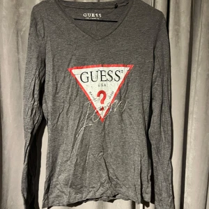 Grå långärmad tröja från Guess, S - Säljer en grå långärmad tröja från Guess med det ikoniska triangellogotypen i rött och vitt på framsidan och text i strass. Tröjan har en v-ringad hals och är perfekt för en avslappnad stil. Passar bra till jeans eller kjol. Använd en gång.