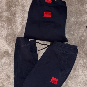 Mjukisset Hugo boss - Säljer en stilren svart sweatshirt och matchande byxor från Hugo. Båda plaggen har en röd logotyp på bröstet respektive benet. Perfekt för en avslappnad look med ribbade muddar och bekväm passform.