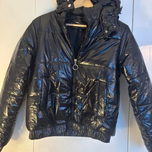 Glossy dunjacka svart. Storlek S-M - Svart glansig jacka med luva. Kort modell. Slutsåld på hemsidan. Storlek M men liten i storlek, passar S. Använd ett fåtal gånger. Nyskick. Djur och rökfritt hem. Nypris 850kr.