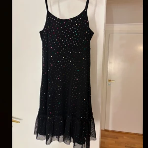 Svart klänning med paljetter - Snygg svart klänning med färgglada paljetter och smala axelband. Den har en volangdetalj längst ner som ger en lekfull touch. Perfekt för en glittrig kväll!