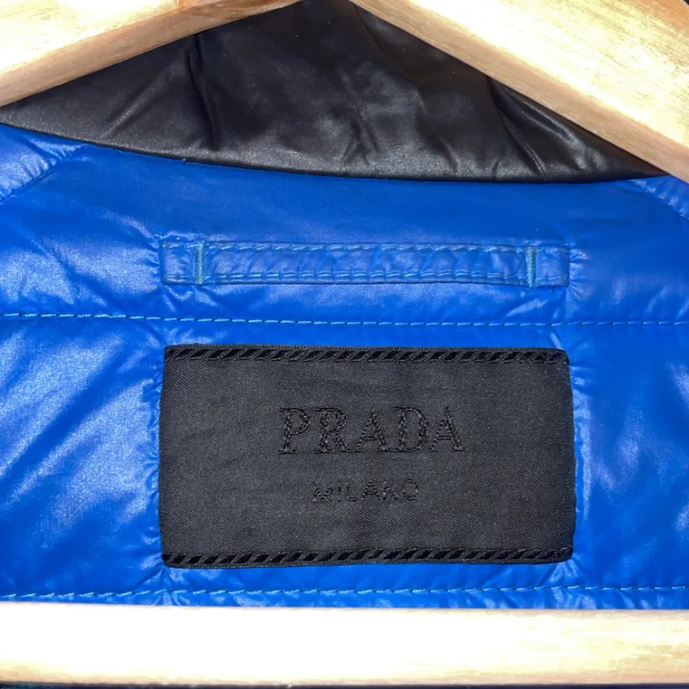 Snygg svart quiltad jacka från Prada med dragkedja och två fickor framtill. Jackan har en stilren design och är perfekt för kyligare dagar. Insidan är fodrad med blått tyg som ger en fin kontrast.. Takit.