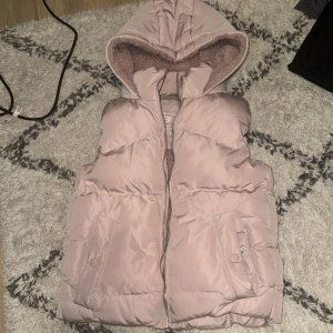 Beige dunväst från FB Sister - Säljer en mysig beige dunväst från FB Sister med huva och dragkedja. Perfekt för kyliga dagar när du vill hålla värmen utan att bära en jacka. Västen har praktiska fickor framtill. Aldrig använd