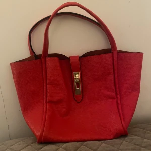 Röd handväska med gulddetalj - Snygg röd handväska i syntetmaterial med en elegant gulddetalj på framsidan. Väskan har två handtag och en rymlig insida med ett mönstrat foder. Perfekt för att ge en färgklick till din outfit.