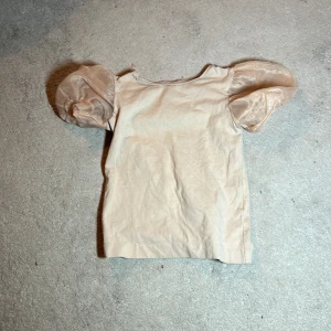 Beige topp med puffärmar från Zara - Säljer en söt beige topp från Zara med puffiga, transparenta ärmar. Perfekt för en stilren och lekfull look. Toppen är i storlek 122 och passar barn i åldern 7 år.