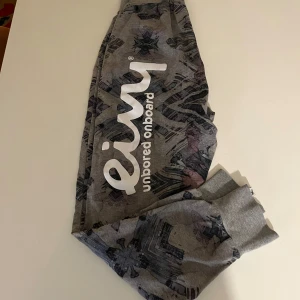 Grå mönstrade mjukisbyxor från Eivy - Säljer ett par grå mjukisbyxor från Eivy med ett unikt mönster i mörkare nyanser. Stort tryck på vänster baksida. Byxorna har en elastisk midja och muddar vid bensluten för extra komfort. Perfekta för en avslappnad stil.