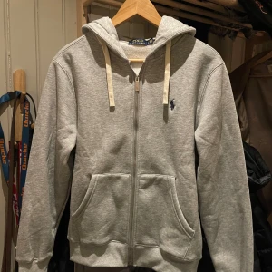 Polo Ralph Lauren zip hoodie - Säljer en populär grå hoodie från Polo Ralph Lauren med dragkedja och klassisk logga på bröstet. Tröjan har långa ärmar och en bekväm huva. Nästintill oanvänd, använd 1 dag, passade inte så säljer den. Storlek M men skulle säga att den är lite mindre i storleken. 