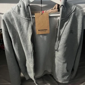 Grå hoodie från Burberry - Säljer en stilren grå hoodie från Burberry med dragkedja och broderad logga på ärmen. Tillverkad i Italien av mjuk bomull med elastan för extra komfort. Perfekt för en avslappnad look. Pris kan diskuteras vid snabb affär och är intresserad av byten.