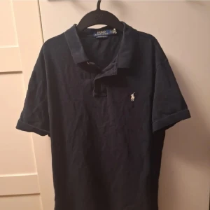 Mörkblå pikétröja från Ralph Lauren - Säljer en stilren mörkblå pikétröja från Ralph Lauren med klassisk krage och korta ärmar. Tröjan har en broderad logotyp på bröstet och är i en custom slim fit passform. Perfekt för en avslappnad men ändå elegant look.