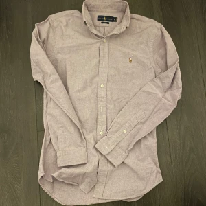 Ralph lauren skjorta - Snygg grålila skjorta från Ralph Lauren i slim fit. Skjortan har långa ärmar och knappar framtill. Den är broderad med det klassiska Ralph Lauren-logotypen på bröstet. Skicket är 9,5/10! Vid övriga frågor eller funderingar är det bara att höra av sig!🙌