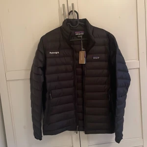 Svart dunjacka från Patagonia - Jackan är helt nytt och har aldrig använts som ni kan se på bilderna är prislappen fortfarande på. Det affärspriset är 2500kr och jag säljer den för 1200kr. Säljer en stilren svart dunjacka från Patagonia med broderad logga och dragkedja framtill. Jackan har en lätt och bekväm passform, perfekt för kyligare dagar. Den är långärmad och har en diskret design som passar till det mesta.