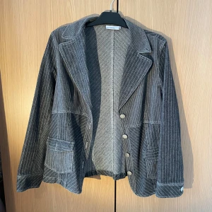 Grå randig kavaj från Almia - Snygg grå kavaj från Almia med smala vita ränder och knappar framtill. Kavajen har en klassisk krage och praktiska fickor. Perfekt för en stilren look.
