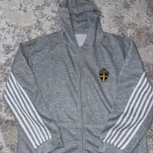 Grå hoodie med dragkedja och emblem - Snygg grå hoodie med dragkedja med byxorna och ett Sverige logo på. Den är helts ny här inte använt mig. Säljer den för denna passade inte mig den står att der är 2XL men den är mindre skulle säga L.