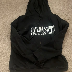 Svart hoodie med Taylor Swift The Eras Tour-tryck - Säljer en svart hoodie med tryck från Taylor Swift The Eras Tour. Den har en stor framficka och justerbar huva med dragsko. På baksidan finns ett färgglatt collage av olika bilder. Perfekt för fans! 🖤
