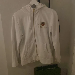 Vit hoodie från Gant - Säljer en stilren vit hoodie från Gant med dragkedja och huva. Tröjan har en broderad logotyp på bröstet och praktiska fickor framtill. Perfekt för en avslappnad stil.