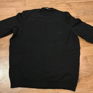 Svart tröja från Hugo Boss - Säljer en stilren svart tröja från Hugo Boss i extrafint merinoull. Tröjor är i storlek L men passar mer som en S/M Tröjan har en diskret logga på bröstet och rund halsringning. Perfekt för en elegant och avslappnad look. Inga fläckar eller slitage. Skriv gärna om ni har frågor allt ifrån pris till fler bilder. 