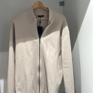 Beige kofta från Massimo Dutti - Snygg beige kofta från Massimo Dutti med dragkedja framtill. Den har en ribbad krage och är perfekt för en stilren look. Passar bra till både vardag och mer uppklädda tillfällen. Storlek XL och aldrig använt den