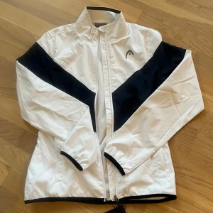 Vit och svart vindjacka från Head - Snygg vit och svart tennis/padel vindjacka från Head med dragkedja och hög krage. Perfekt för alla sportiga aktiviteter. Jackan har långa ärmar och elastiska muddar för en bekväm passform. Den är dam men skulle lika gärna kunna vara unisex (Nypris 800kr) skick: helt ny. 