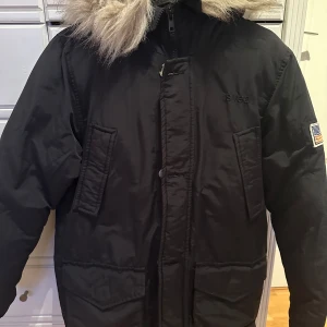 Svart parkajacka med pälsdetalj - Snygg svart parkajacka från Svea med pälsdetalj på huvan. Jackan har en dragkedja framtill och praktiska fickor. 