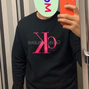 Svart sweatshirt från Calvin Klein - Snygg svart sweatshirt från Calvin Klein med rosa CK-logga på bröstet. Tröjan har långa ärmar och en klassisk rund halsringning. Perfekt för en stilren och avslappnad look. Endast använd 2 gånger.