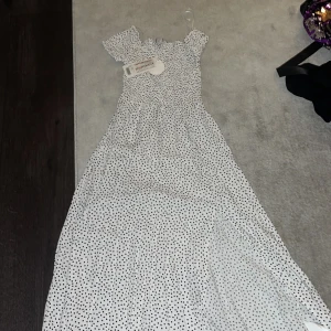 Vit prickig långklänning - Helt oanvänd med prislapp! Charmig vit långklänning med svarta prickar och korta ärmar. Perfekt för somriga dagar med sin luftiga design och bekväma passform. Klänningen har en söt offshoulder-stil som ger en romantisk touch.