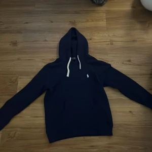 Mörkblå hoodie från Polo Ralph Lauren - Säljer en snygg mörkblå hoodie från Polo Ralph Lauren med klassisk logga på bröstet. Tröjan har en justerbar huva med vita snören och en stor ficka framtill. Perfekt för en avslappnad stil. Ny pris 2200. Mitt pris 600. Pris kan diskuteras vid snabb affär.