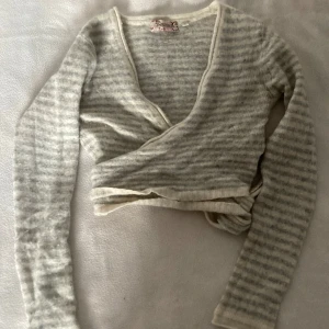Grå och vit randig omlottopp från Indiska - Säljer en mysig grå och vit randig omlottopp från Indiska. Toppen har långa ärmar och knytband framtill för en justerbar passform. Perfekt för en avslappnad stil.