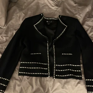 Svart kavaj från Carole Little - Elegant svart kavaj från Carole Little med vita detaljer. Kavajen har en stilren design med vita kantband längs kanterna och fickorna. Kavajen är helt ny förutom att en tråd pickar ut. 