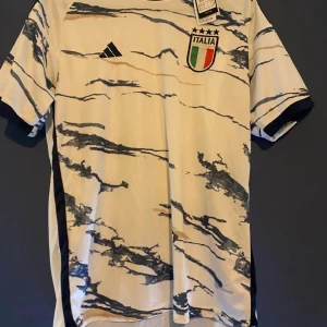 Italien fotbollströja från Adidas - Snygg fotbollströja med blått mönster och Italiens emblem på bröstet. Tröjan har korta ärmar och en modern design med Adidas-logga. Perfekt för fotbollsfans som vill visa sitt stöd för Italien.
