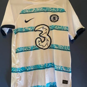 Vit och blå Chelsea fotbollströja - Säljer en vit Chelsea fotbollströja från Nike med blå ränder och klubbens emblem på bröstet. Tröjan har korta ärmar och ett stilrent mönster i ränderna. Perfekt för fans som vill visa sitt stöd.