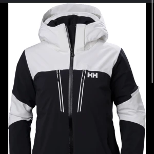 Svart och vit jacka från Helly Hansen - Snygg svart och vit skid jacka från Helly Hansen med dragkedja och huva. Perfekt för kyliga dagar med sin funktionella design och stilrena look. Orginalpis 3500, säljer för 2500. Bra skick
