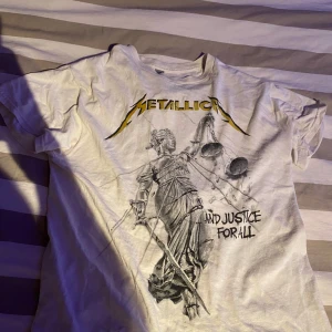 Vit Metallica t-shirt - Cool vit t-shirt med Metallica-tryck fram och bak. Framsidan har en illustration av Lady Justice och texten 'And Justice For All'. Baksidan visar bandmedlemmarnas ansikten och texten 'Hammer of Justice Crushes You'. Perfekt för fans av bandet!