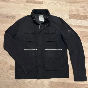 Svart jacka från J.Lindeberg - Snygg svart jacka från J.Lindeberg med dragkedja och knappar. Perfekt för vår och höst. 