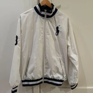 Vit vindjacka från Ralph Lauren - Snygg vit vindjacka från Ralph Lauren med dragkedja och broderad logga på bröstet. Jackan har långa ärmar och ribbade muddar i svart och vitt vid ärmslut och nederkant. Perfekt för en stilren look.